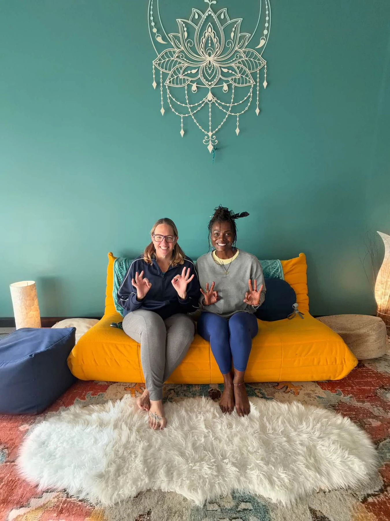 Two smiling people seated side by side on the yellow sofa at Calm Soul Studio.